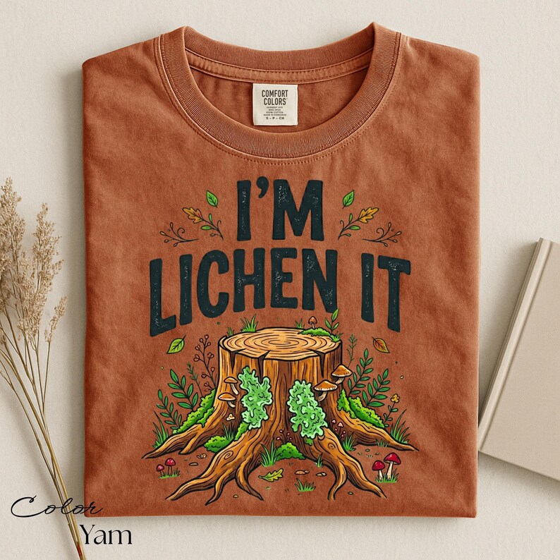 Puede incluir: Una camiseta de color &oacute;xido con la frase "I'M LICHEN IT" en letras negras. El gr&aacute;fico presenta un toc&oacute;n de &aacute;rbol con l&iacute;quenes verdes, setas y follaje. La camiseta est&aacute; doblada y tiene una etiqueta en el centro superior.