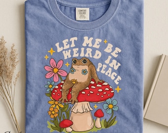 Op de afbeelding: Een jeansblauw t-shirt met een afbeelding van een bruine kikker die op een grote rood-witte paddenstoel zit. De tekst "LET ME BE WEIRD IN PEACE" staat erboven gedrukt, omringd door kleurrijke bloemen en gele sterren.