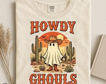 Op de afbeelding: Een ivoorkleurig t-shirt met een afbeelding van een geest die een bruine cowboyhoed en laarzen draagt, staand in een woestijnlandschap met cactussen en een zonsondergang. De tekst HOWDY GHOULS is in oranje gedrukt in een western-lettertype.