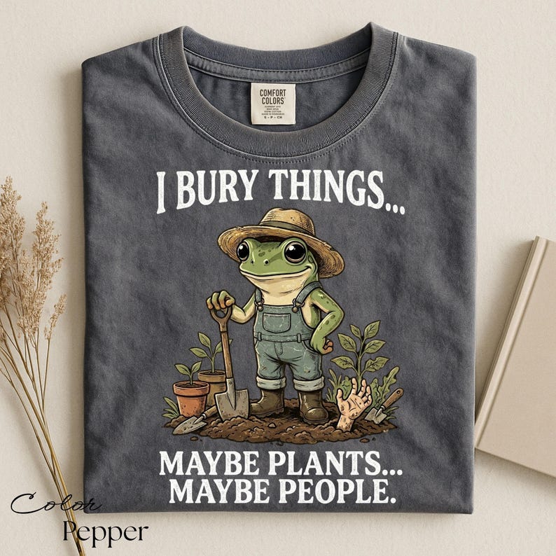 Może przedstawiać: Ciemnoszary t-shirt z grafiką żaby w słomkowym kapeluszu i ogrodniczkach, trzymającej łopatę w ogrodzie. Napis głosi I BURY THINGS... MAYBE PLANTS... MAYBE PEOPLE. Koszulka posiada metkę marki Comfort Colors przy kołnierzu.