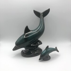 May include: A pair of ceramic dolphin figurines. The larger dolphin is dark green and black with a glossy finish. The smaller dolphin is a lighter shade of green. Both dolphins are standing on a black base.