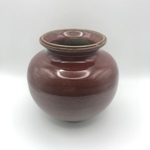May include: A round, red ceramic vase with a slightly flared rim. The vase has a glossy finish and a subtle, uneven texture.
