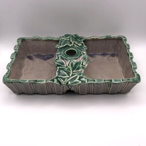 May include: A green and purple ceramic planter with two rectangular compartments and a central hole. The planter has a leafy design around the hole and a textured, ribbed design around the edges.