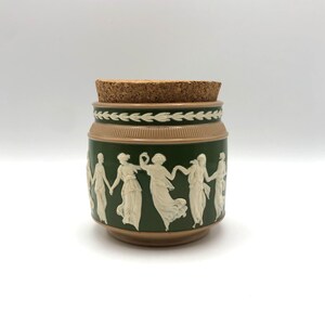 May include: A green and brown ceramic jar with a cork lid. The jar features a white relief design of figures dancing around the side.