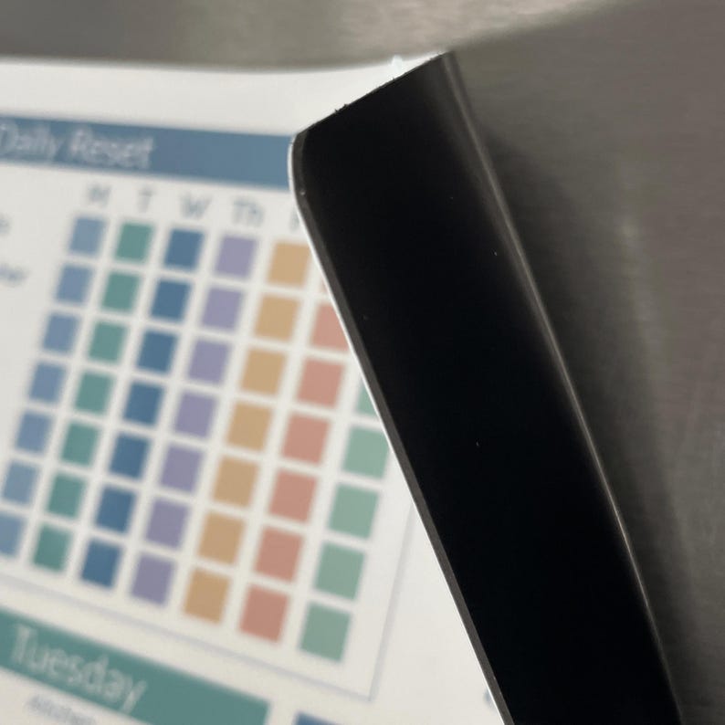 May include: A corner view of a white and black magnetic calendar. The calendar has a grid of squares in various colours, with the words "Daily Reset" and "Tuesday" visible. A black magnetic sheet is partially covering the calendar.