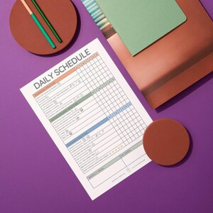 May include: A printed daily schedule on a white paper with orange, green, and blue highlights. The schedule is on a purple surface with a brown coaster, a green notebook, and two pencils.
