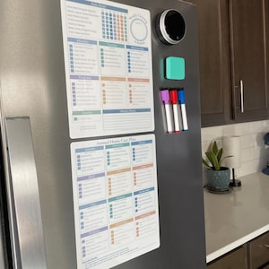 May include: A stainless steel fridge door features a magnetic weekly cleaning schedule, a daily reset checklist, and an annual home care plan. A smart home device, a small teal square, and three coloured dry-erase markers are also attached.