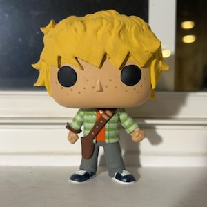 May include: A custom-made pop figure of a young person with yellow hair, wearing a green and orange plaid shirt, grey pants, and brown boots. The figure has a brown leather belt and a brown satchel.