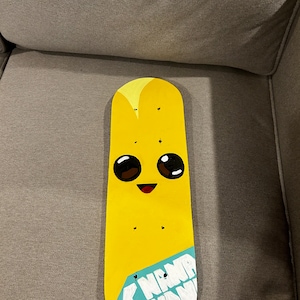 Pu&ograve; includere: Un mazzo di skateboard giallo dipinto con un viso di banana dei cartoni animati e le parole "Nana Nana" su un'etichetta blu.