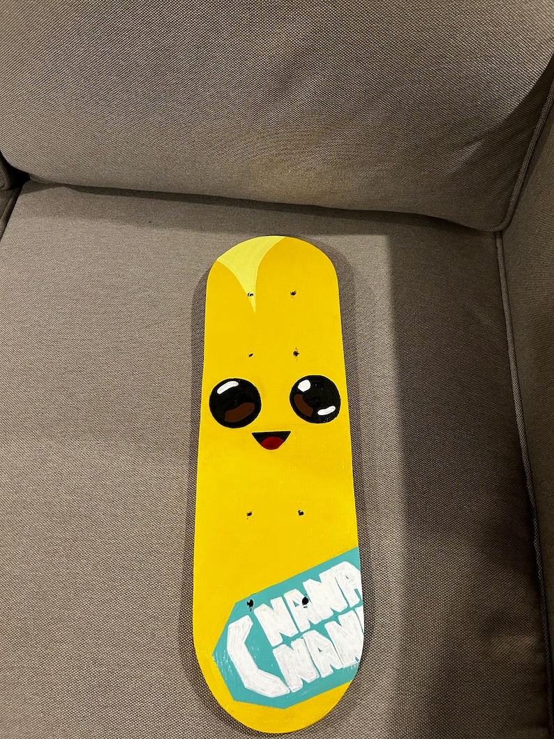 Pu&ograve; includere: Un mazzo di skateboard giallo dipinto con un viso di banana dei cartoni animati e le parole "Nana Nana" su un'etichetta blu.