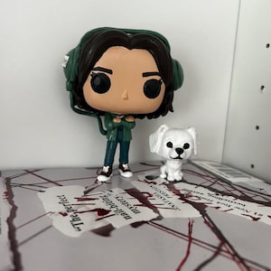 以下が含まれることがあります： 緑と黒のポップビニールフィギュアで、ヘッドホンと緑色のジャケットを着た女性が描かれています。フィギュアは赤い印のついた本の上に立っています。女性の隣には白い犬のフィギュアが立っています。