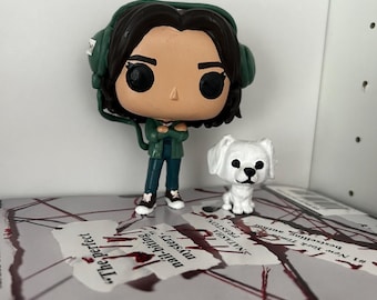 以下が含まれることがあります： 緑と黒のポップビニールフィギュアで、ヘッドホンと緑色のジャケットを着た女性が描かれています。フィギュアは赤い印のついた本の上に立っています。女性の隣には白い犬のフィギュアが立っています。