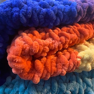 May include: Close-up of a chunky knit blanket in shades of blue, orange, and yellow. The soft, textured yarn is arranged in horizontal layers, creating a cozy and inviting appearance. The blanket appears to be hand-knitted.