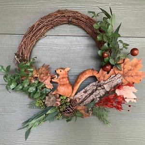 May include: A rustic grapevine wreath decorated with a carved wooden squirrel, fall leaves, pinecones, and berries. The wreath features a variety of textures and colors, including brown, orange, green, and red. Perfect for autumn decor.