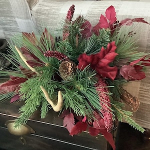 May include: A decorative centerpiece featuring evergreen branches, red leaves, and berry clusters. Accents include pine cones and small antler pieces, creating a rustic, seasonal arrangement. The arrangement is placed on a dark surface.