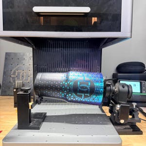 May include: A tumbler with a geometric design in shades of blue, teal, and black is positioned within a laser engraving machine. The tumbler is held in place by a series of clamps and supports, ready for the engraving process.