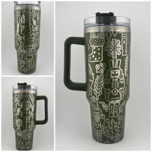 May include: A green stainless steel travel mug with a white design of a nutcracker, Christmas trees, and other holiday imagery. The mug has a lid and a handle.