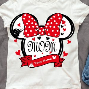Puede incluir: Camiseta blanca con una ilustración en blanco y negro de las orejas de Minnie Mouse con un lazo rojo con lunares blancos. El texto "Mom" está dentro de las orejas y "Your Name" está en una pancarta roja debajo.
