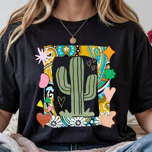 Puede incluir: Camiseta negra con un gráfico colorido de un cactus verde dentro de un marco decorativo. El marco incluye corazones, estrellas y patrones florales en tonos rosa, amarillo y azul. La camiseta está hecha de un material suave.