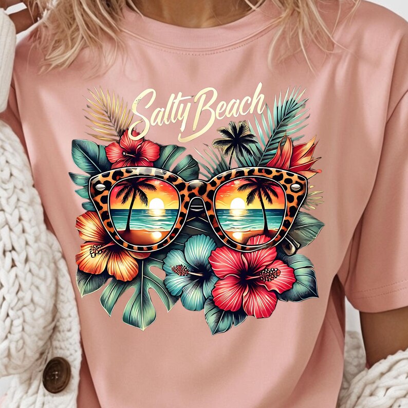 May include: A pink t-shirt with a graphic print of sunglasses with a tropical beach scene reflected in the lenses. The sunglasses are surrounded by tropical flowers and leaves. The text "Salty Beach" is printed in gold above the sunglasses.