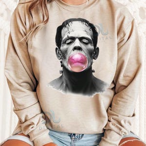 Op de afbeelding: Een beige sweatshirt met een zwart-wit afbeelding van Frankenstein die een roze bel blaast. De afbeelding is gecentreerd op het sweatshirt.