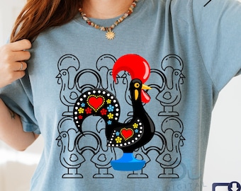 May include: A light blue t-shirt featuring a stylized black rooster with a red comb and heart-shaped accents. The rooster is surrounded by a pattern of rooster outlines. The shirt has a crew neck and short sleeves.