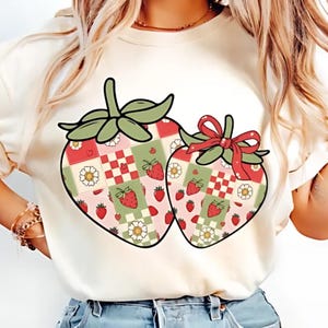 May include: A cream-colored t-shirt featuring two large strawberry designs. Each strawberry has a green stem and leaf design. The strawberries are decorated with a patchwork pattern of red, green, and white squares, strawberries, and flowers.