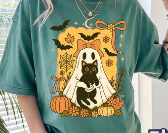 May include: Green t-shirt featuring a Halloween-themed graphic. The design includes a ghost holding a black cat, surrounded by bats, pumpkins, and fall foliage. A large orange bow is at the top of the design.