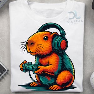以下が含まれることがあります： ヘッドフォンとゲームコントローラーを持ったカピバラのカラフルなイラストが描かれた白いTシャツ。カピバラはオレンジ、ティール、赤色です。ゲーマーにぴったりの遊び心のあるデザインのシャツです。