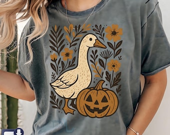 May include: A gray t-shirt featuring a cream-colored goose, a jack-o'-lantern, and floral elements in brown and yellow. The design is centered on the shirt, creating a fall or Halloween theme.