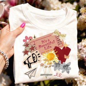 Puede incluir: Camiseta blanca con un diseño de collage con el texto "It's a Beautiful Life". El diseño incluye un megáfono, estrellas, flores, un corazón, un sol y un avión de papel. La camiseta la sostiene una persona.