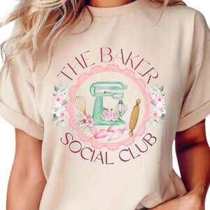 May include: A beige t-shirt with a pink and white floral design and a green stand mixer. The text "The Baker Social Club" is printed in burgundy on the shirt.