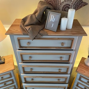 Op de afbeelding: Een lichtblauwe en houten commode met vijf lades, elk omlijnd in goud. De bovenkant is van hout en de knoppen zijn rond. Een ingelijste print, kaarsen en een decoratief kussen liggen bovenop. Twee bijpassende nachtkastjes staan aan weerszijden.