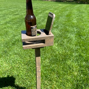 Pode incluir: Um suporte de madeira com acabamento marrom, com um suporte de telefone integrado e um porta-copos para uma garrafa. O suporte é montado em uma estaca de madeira e é colocado em uma área gramada.