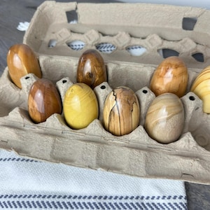 Puede incluir: Ocho huevos de madera en varios tonos de marrón y amarillo están dispuestos en una caja de huevos. Los huevos son lisos y tienen un patrón de veta de madera natural.