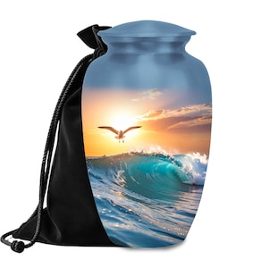 May include: A blue cremation urn with a scenic ocean wave and seagull design. The urn is paired with a black drawstring bag. The image on the urn features a vibrant sunset over the ocean, with a seagull in flight.