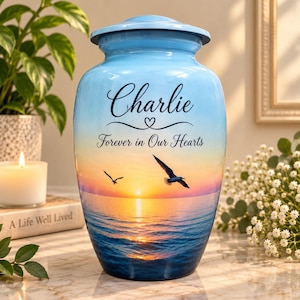 May include: A light blue cremation urn featuring a sunset ocean scene with two flying seagulls. The text "Charlie" is printed in black script above the phrase "Forever in Our Hearts" in a smaller font. The urn has a matching lid.