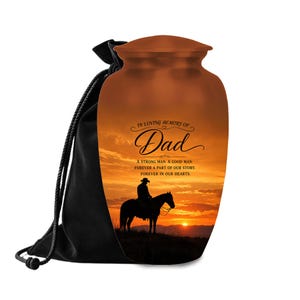 May include: A copper-colored metal cremation urn featuring a sunset landscape with a silhouette of a person on horseback. The text reads: IN LOVING MEMORY OF Dad. A STRONG MAN. A GOOD MAN. FOREVER A PART OF OUR STORY. FOREVER IN OUR HEARTS. Includes a black bag.