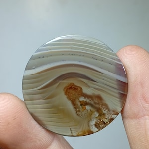 May include: A polished, round agate stone with a layered pattern of beige, white, and brown hues. The stone features a unique, organic pattern with a central, rust-colored inclusion. The stone is held between fingers.