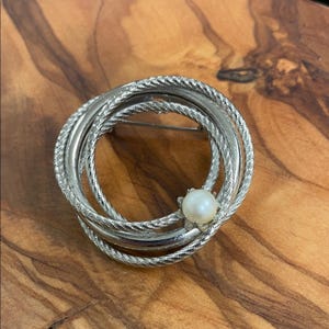 May include: Silver-toned brooch with three interlocking rings and a central pearl accent. The rings have a textured, rope-like design. The brooch is set against a wooden background, showcasing its elegant design.