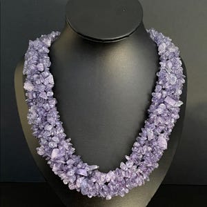 May include: A multi-strand necklace composed of small, irregular, lavender-colored gemstones. The necklace is displayed on a black bust, showcasing its layered design and the varying shades of purple within the stones. The necklace is a statement piece.