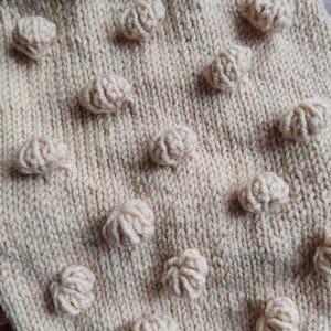 Op de afbeelding: Close-up van een crèmekleurige gebreide stof met verhoogde, getextureerde bobbles. De bobbles zijn gelijkmatig verdeeld over het oppervlak, waardoor een tastbaar, decoratief patroon ontstaat. Het breiwerk ziet er zacht en gezellig uit, geschikt voor een sjaal of deken.