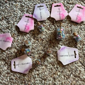 May include: A collection of necklaces featuring small glass bottles filled with colorful beads and charms. Each necklace has a silver chain and is displayed with a tag that reads "Abby's Treasures". The tags have a pink and purple watercolor design.