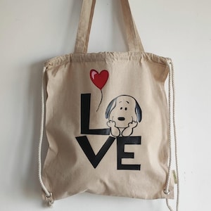 Könnte beinhalten: Eine beige Canvas-Tragetasche mit Kordelzug. Die Tasche zeigt eine Schwarzweiß-Illustration von Snoopy, der einen roten Herzluftballon hält. Der Text "LOVE" ist in schwarzen Buchstaben auf der Tasche aufgedruckt.