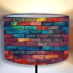 Pode incluir: Um abajur com um padrão de tijolos colorido. O design apresenta uma variedade de cores, incluindo vermelho, azul, verde e amarelo, dispostas em um design de parede de tijolos. O abajur é cilíndrico com um interior branco.