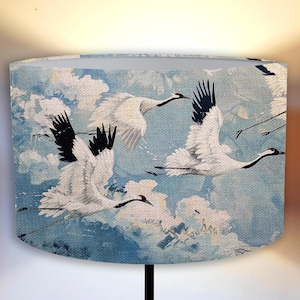 May include: A lampshade featuring a blue background with a white and black crane design. The cranes are flying in formation against a cloudy sky.