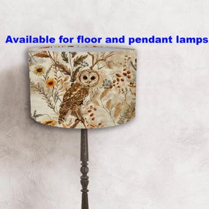 Könnte beinhalten: Ein Lampenschirm mit beigefarbenem Hintergrund und einer braunen Eule mit weißem Gesicht und gelben Augen. Die Eule ist von braunen Ästen und Blättern umgeben, und kleine gelbe Blumen sind über das gesamte Design verteilt.