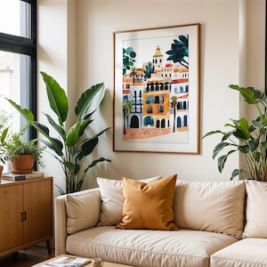 May include: A white sofa with a mustard yellow cushion in front of a framed print of a colourful illustration of buildings. The print is in a wooden frame and hangs on a white wall. There are two large green plants in the room.