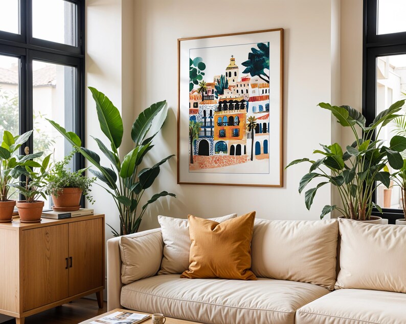 May include: A white sofa with a mustard yellow cushion in front of a framed print of a colourful illustration of buildings. The print is in a wooden frame and hangs on a white wall. There are two large green plants in the room.