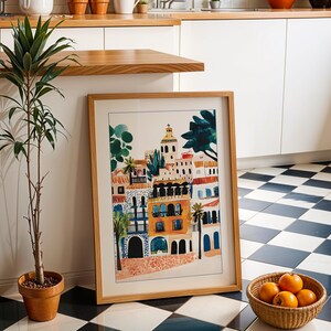 May include: A framed print of a colourful illustration depicting a cityscape with buildings, trees, and a blue sky. The print is in a wooden frame and is hanging on a white wall.